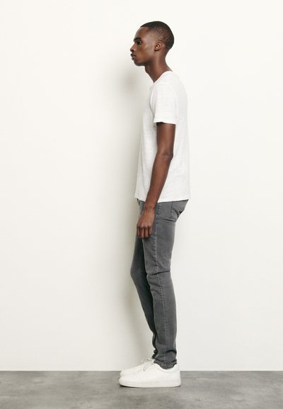 Lys grå t-skjorte med kort erm, slim-fit mørkegrå jeans og hvite sneakers; sidevinkel viser et enkelt og moderne utseende.