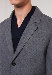 Chaqueta de mezcla de lana gris con cuello con muescas, cierre de un solo botón negro y detalles de costura visibles, superpuesta sobre un suéter azul marino.
