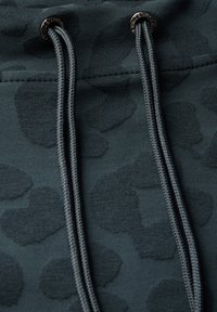 Tejido de sudadera de color verde oscuro con un sutil patrón de leopardo. Presenta cordones resistentes con terminales metálicos. Textura suave en general.