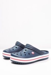 Zoccoli blu navy con fori di ventilazione, suola a strisce bianche e rosse, materiale plastico morbido, cinturino regolabile e dettaglio del logo di lato.