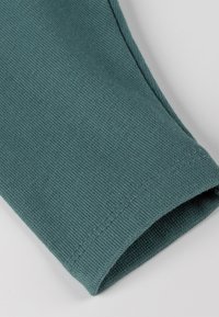 Nahaufnahme des Ärmelsaums eines türkisfarbenen gerippten Kleidungsstücks, das den strukturierten Stoff und die sauberen Nähte vor einem hellen Hintergrund zeigt.