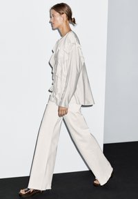 Light gray ruffled blouse paired with wide-leg white trousers. The ensemble features a relaxed fit with smooth textures and minimal accents.