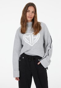 Grauer Strickpullover mit einem weißen Herz-Logo und gerippter Textur. Lange Ärmel mit marineblauen Streifen und weiten Bündchen. Kombiniert mit schwarzen Hosen.