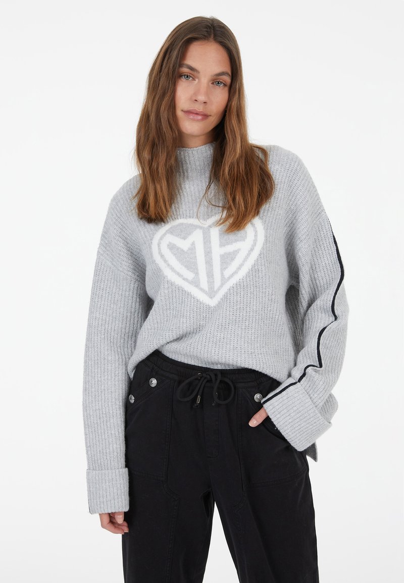 Grauer Strickpullover mit einem weißen Herz-Logo und gerippter Textur. Lange Ärmel mit marineblauen Streifen und weiten Bündchen. Kombiniert mit schwarzen Hosen.