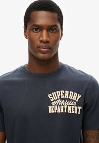 Marinblå bomulls t-shirt med en broderad "SUPERDRY Athletic Department" logga i beige på bröstet, med en klassisk rund halsringning.