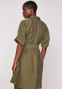 Donna con capelli corti che indossa orecchini a cerchio dorati e un vestito verde oliva con cintura e maniche arrotolate, di profilo.