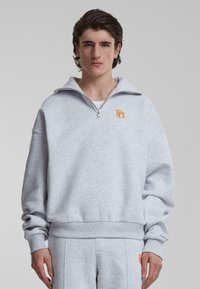 Lys grå hettegenser med høy krage, halvlukking, oransje brodert logo på brystet, og elastiske mansjetter og midje, laget av mykt stoff.