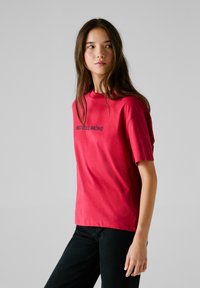 Röd bomullströja med korta ärmar, med texten "RED BULL RACING" i blått. T-shirten har rund hals och en avslappnad passform.