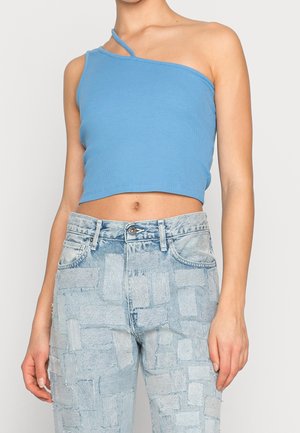 Frau trägt ein hellblaues, ein-schulteriges Crop-Top und hoch taillierte, hell gewaschene Jeans mit Patchwork-Details.