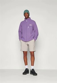 Mies, joka pukeutuu vihreään neulottuun myssyyn, purppuraan vetoketjulliseen takkiin, vaaleisiin shortseihin, valkoisiin sukkiin ja mustiin lenkkareihin, seisoo kädet taskuissa.