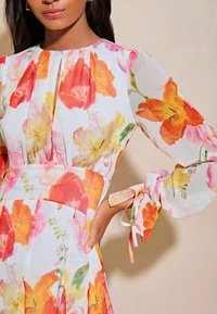Rochie florală dintr-un material ușor, transparent, cu o varietate de flori colorate în roșu, portocaliu și galben. Mâneci lungi cu accente legate.