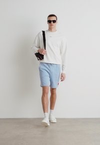 Hanorac alb, pantaloni scurți bleu deschis, geantă crossbody neagră, ochelari de soare negri și adidași albi. Texturi fine, croială lejeră.