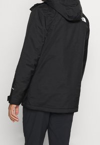 Veste noire avec une finition texturée, capuche ajustable et poignets élastiques. Détail du logo au dos. Design simple et fonctionnel.