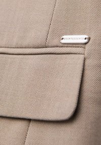 Gros plan d'un tissu chevron beige avec un rabat et une petite étiquette métallique gravée du mot "QUINTESSENCE."