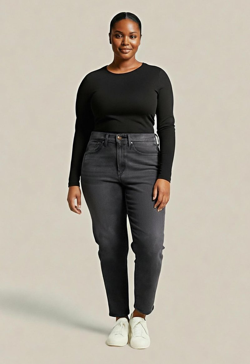 Pewna siebie kobieta stoi w czarnej bluzce z długim rękawem, ciemnoszarych spodniach z wysokim stanem i białych sneakersach na jasnym tle.