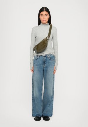 Woman standing facing forward wearing a grey long-sleeve top, wide-leg blue jeans, black shoes, and an olive green crossbody bag.