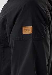 Svart jackärm med ett brunt rektangulärt märke märkt "Swedemount" med en bergslogotyp.