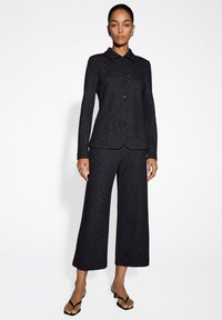 Completo nero con giacca colletto e chiusura con bottoni, abbinata a pantaloni a gamba larga. Entrambi i pezzi presentano un delicato effetto metallico.