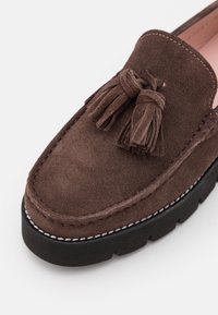 Mocassin en daim marron avec pompons, bout arrondi, surpiqûres blanches contrastantes et semelle extérieure épaisse en caoutchouc noir.