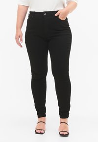 Jeans skinny negros de denim, con una textura suave, talle alto, bolsillos delanteros y cierre de botones. Combinados con sandalias negras de tiras.