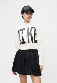 Donna che indossa un maglione bianco con cerniera e la scritta nera "LIKE" in grande, pantaloncini neri imbottiti e un cappellino da baseball in pelle nera, in piedi contro uno sfondo bianco.