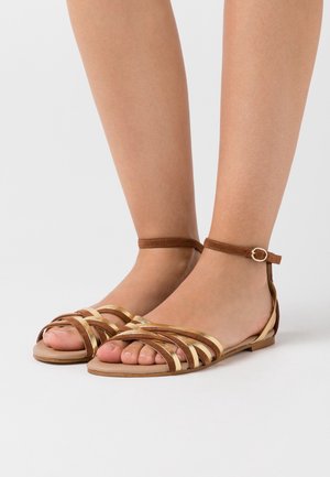 Brązowe i złote sandały na płaskiej podeszwie z paskiem wokół kostki oraz plecionym wzorem na palcach. Gładka faktura, minimalistyczny styl.
