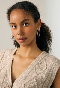 Bež kudumine vest V-kaelusega ja tekstuuriga nöörmustriga, mille juurde kuuluvad kuldsed rõngaskõrvarõngad. Loomulikud lokkis juuksed raamistavad nägu.