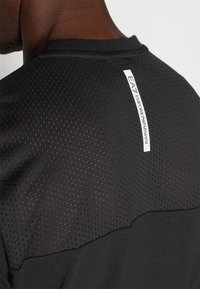 Svart träningströja med en texturerad mesh-övre del, slät nedre del och en märkesetikett på baksidan av kragen.