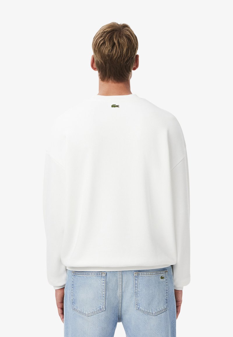 Felpa bianca con vestibilità loose, caratterizzata da un colletto rotondo e un piccolo logo verde sulla parte superiore della schiena. Abbinata a jeans azzurri chiari.