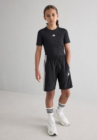 Melna sporta krekls ar baltu logo, apvienots ar melniem šortiem, kuriem ir baltas sānu svītras. Modelis valkā baltas kurpes un svītrainas zeķes.