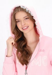 Sudadera con cremallera rosa con capucha adornada con encaje, con letras blancas bordadas y un pequeño logo de corazón bordado. Tela suave, diseño casual.