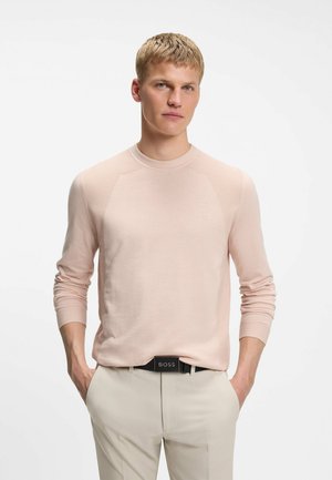 Junger Mann mit kurzen blonden Haaren, der einen hellrosa Langarmpullover und beige Hosen mit einem schwarzen BOSS-Gürtel trägt, Hände in den Taschen.