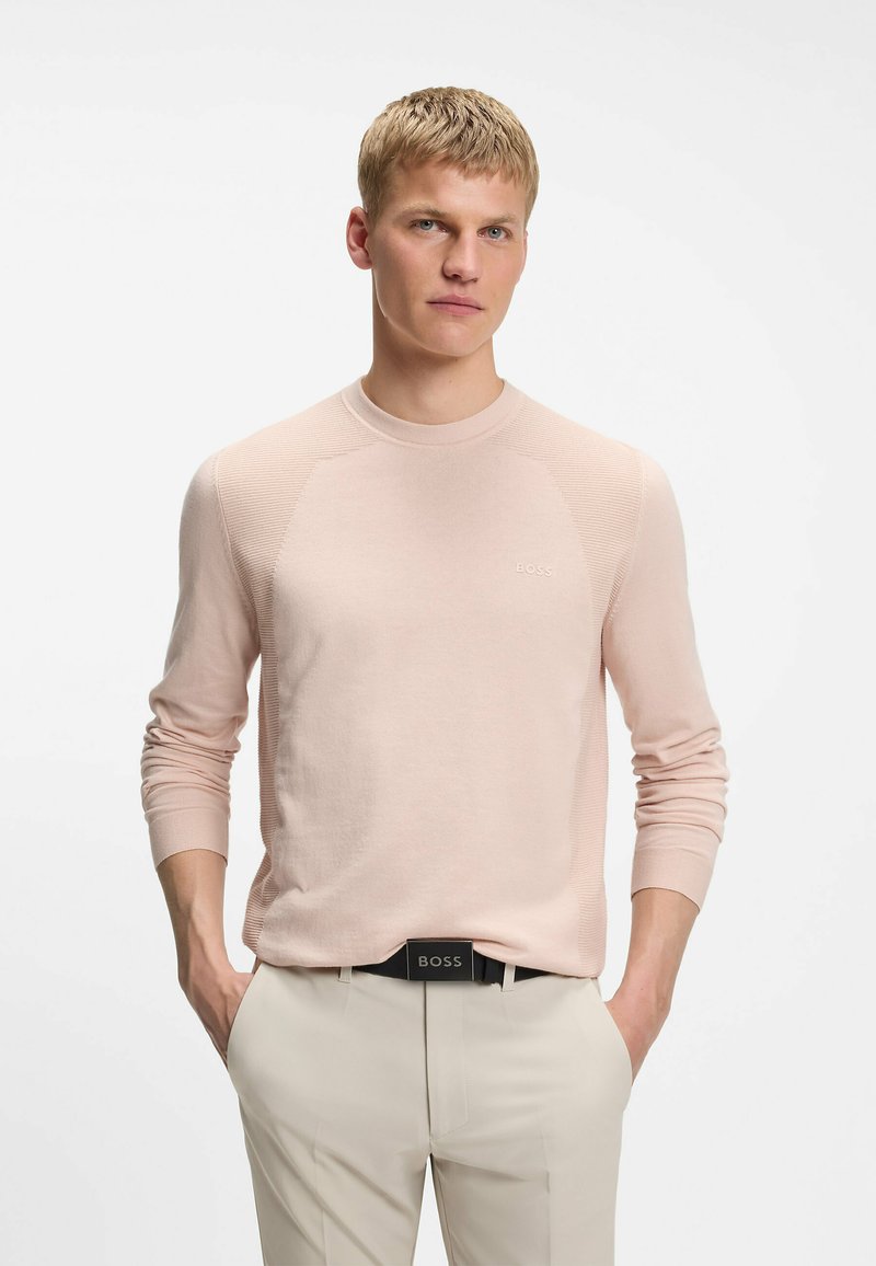 Jeune homme aux cheveux blonds courts portant un pull léger rose à manches longues et un pantalon beige avec une ceinture noire BOSS, les mains dans les poches.