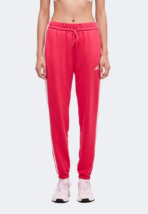 Femme portant un pantalon de survêtement rose Adidas avec des bandes blanches sur les côtés et des baskets rose clair, debout sur un fond uni.