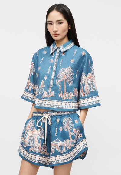 Νέα γυναίκα που φοράει ταιριαστό μπλε μπλουζάκι με κοντά μανίκια και σορτς με κορδόνι στη μέση και διακοσμητικά μοτίβα με ήλιο και φοίνικες.
