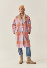 Roosa ja oranžika tooniga ruuduline villane mantel, millel on sälgkragen, kaks esimest taskut ja lilleline tikand sinistel teksadel, millele on kombineeritud pahkluu saapad.