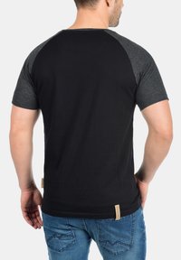 Black short-sleeve T-shirt with gray raglan sleeves, cotton material, straight cut, visible stitching, and small brand tag at the lower side.