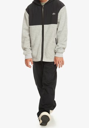 Bluza z kapturem na zamek błyskawiczny z szarym korpusem, czarnym kapturem oraz kontrastowymi poziomymi panelami; do niej czarne spodnie i jasne trampki.