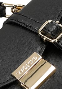 Sac en cuir noir avec des accessoires en métal doré, comprenant une boucle et une plaque logo. Texture lisse et design structuré avec une bandoulière.