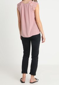 Světle růžový top s krajkovými krátkými rukávy, plisovaná zadní část, spárovaný s černými černými džíny s ohrnutými nohavicemi a černými sandály, pohled zezadu.