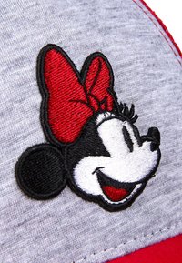 Gorra gris con un personaje bordado en blanco y negro con un lazo rojo, sobre un fondo de tela texturizada, con acentos rojos en contraste.