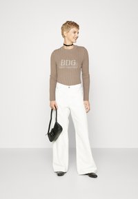 Beige stripet strikkegenser med logo, paret med hvite vide bukser. Modellen holder en liten svart skulderveske. Jevn, avslappet silhuett.