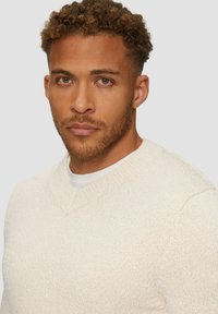 Gebreide trui in licht roomkleur met een subtiele structuurpatroon. Heeft een V-hals en geribde manchetten, gelaagd over een wit T-shirt.