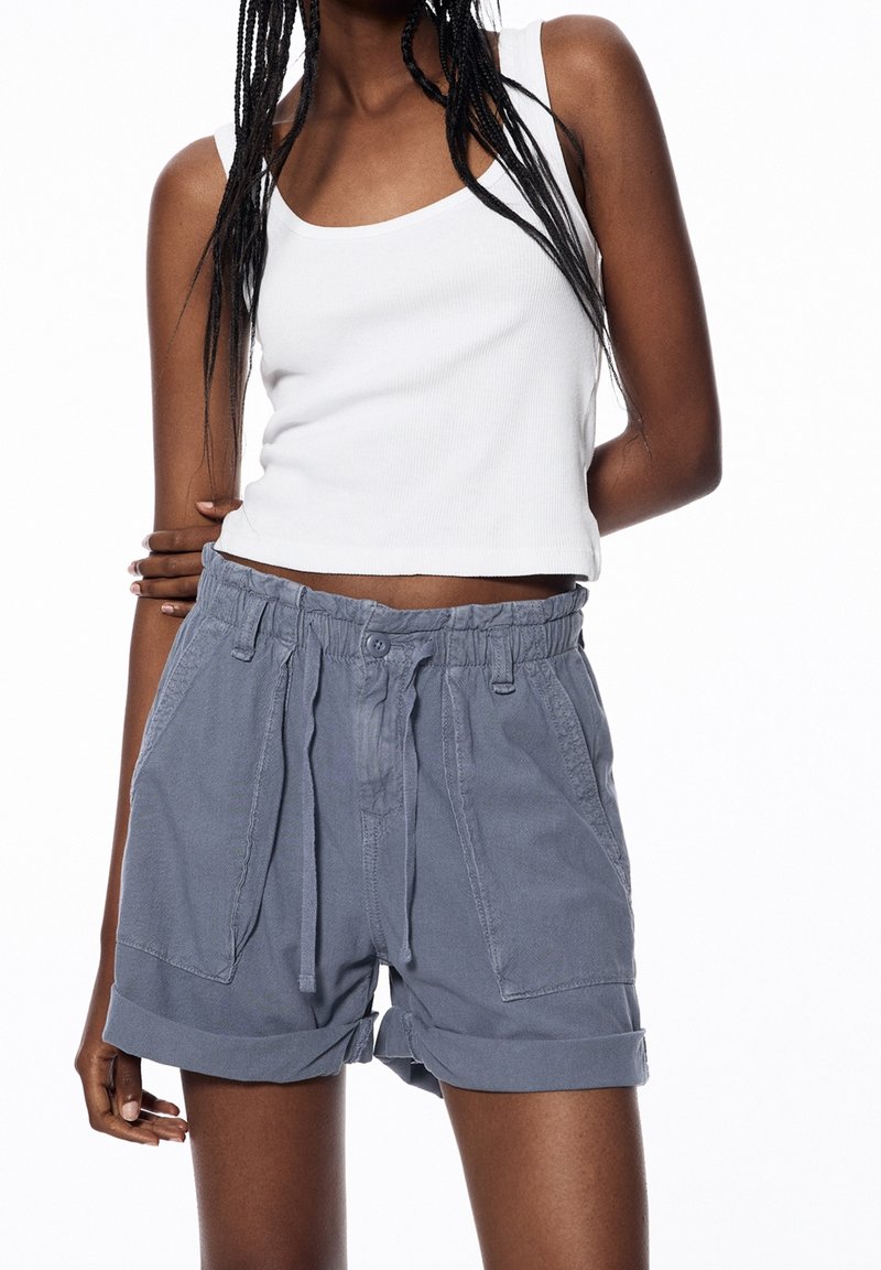 Una donna indossa un top bianco senza maniche e pantaloni corti blu a vita alta con coulisse, tasche frontali e orli arrotolati.