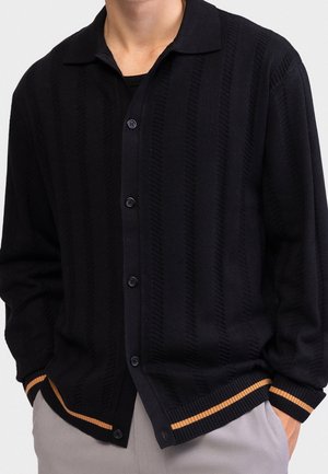 Homme portant un cardigan noir boutonné avec des motifs verticaux subtils et des poignets rayés orange, associé à un pantalon gris clair.
