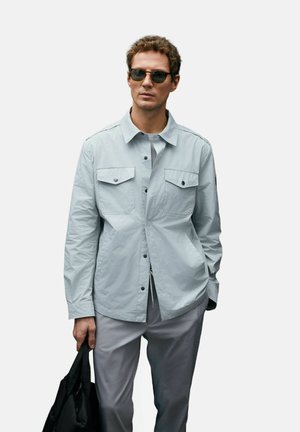 Mann trägt hellgraue, geknöpfte Jacke, passende Hose, dunkle Sonnenbrille, hält schwarze Tasche in der linken Hand vor einfarbigem Hintergrund.