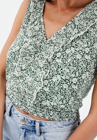 Haut court vert à motif floral blanc, col volanté et taille smockée. Tissu léger, manches courtes et col en V.