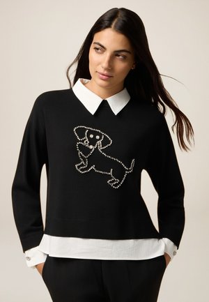 Zwarte trui met een witte shirtkraag, versierd met rhinestones die een hondenontwerp vormen. De onderste zoom heeft een gelaagde witte stof.
