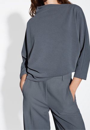 Vrouw draagt een losvallende donkergrijze top met driekwart mouwen en bijpassende hoge taillebroek, handen in de zakken, staand tegen een witte achtergrond.