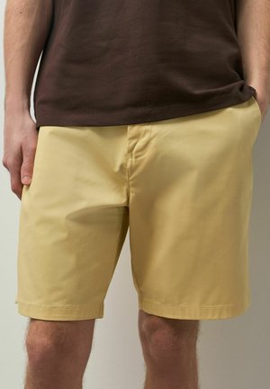 Mann trägt beige knielange Shorts und ein dunkelbraunes Hemd, steht mit einer Hand in der Tasche vor einem schlichten Hintergrund.